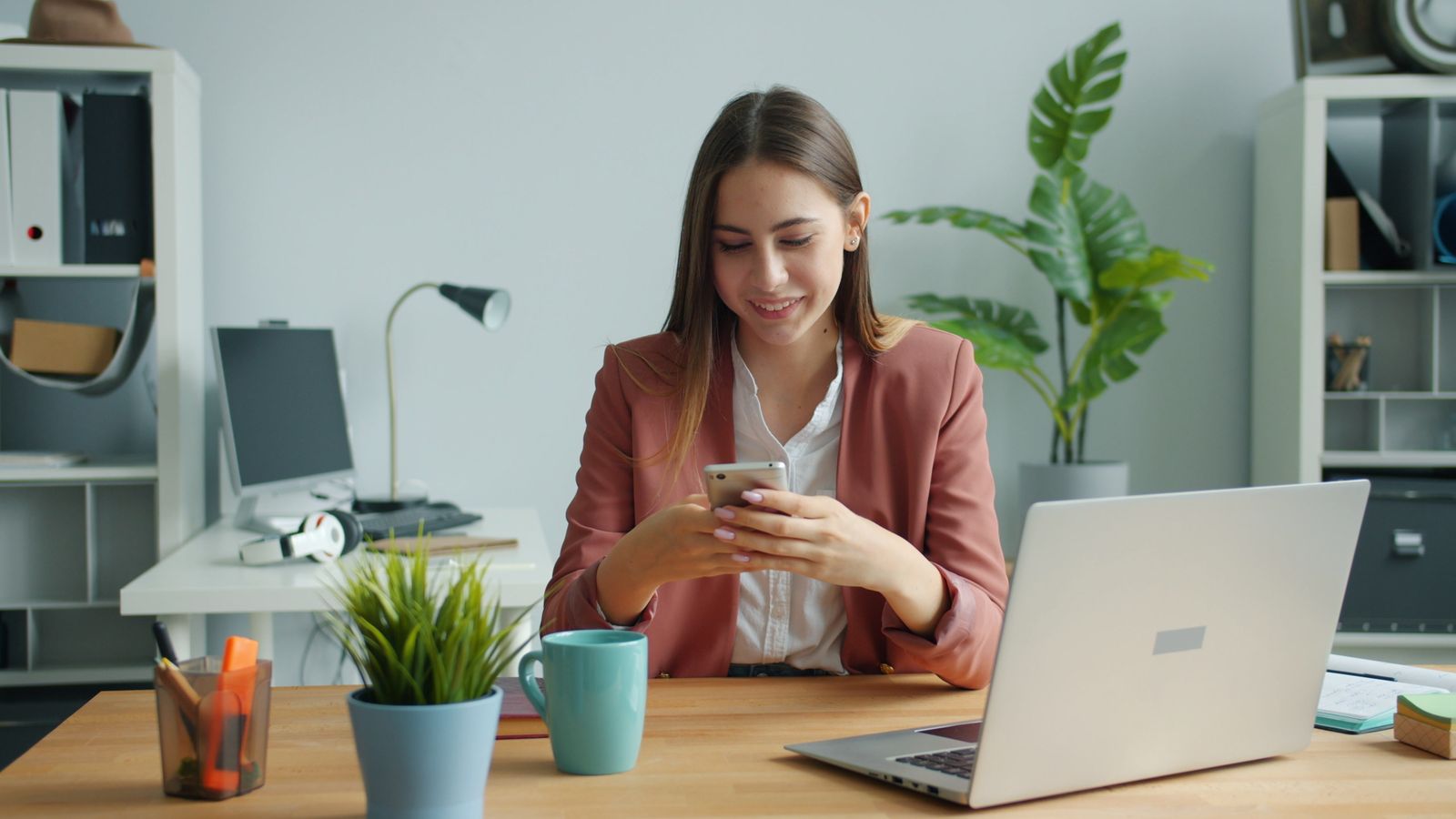 A smiling businesswoman looks at a smartphone at an office desk